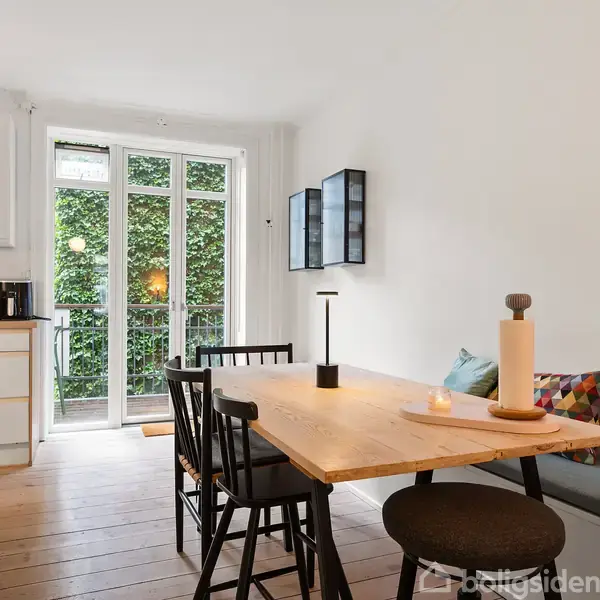 Træbord med sorte stole og lysestage i en lys, moderne spisestue. Til venstre ses et køkkenområde med kaffemaskine. Store vinduer i baggrunden viser en grøn udsigt.
