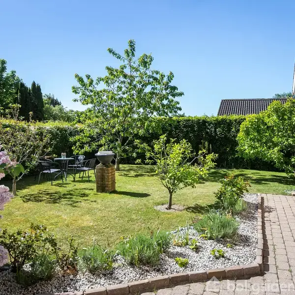 Have en havegrill står midt på en velplejet græsplæne omgivet af buske og blomsterbede. Et cafébord med stole ses under træet, og stien fører langs huset.