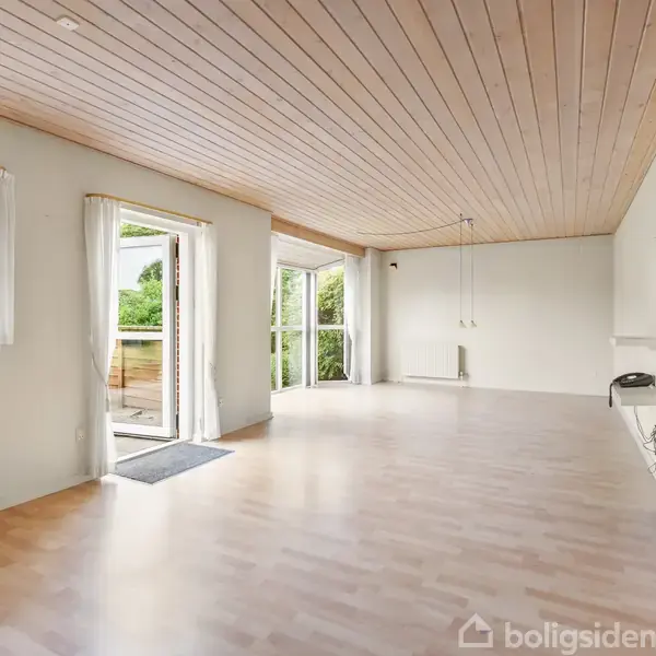Stue med lyst trægulv og træloft, åbne terrassedøre ud til have. Store vinduer lader naturligt lys ind, gardiner trukket til siden. Radiator står under vinduet.