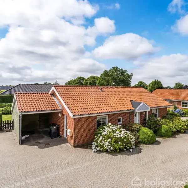 Rødt murstenshus med rødt tegltag og carport parkeret ved siden af. Området har en brostensbelagt indkørsel, omgivet af grønne buske og græs med flere lignende huse bagved.