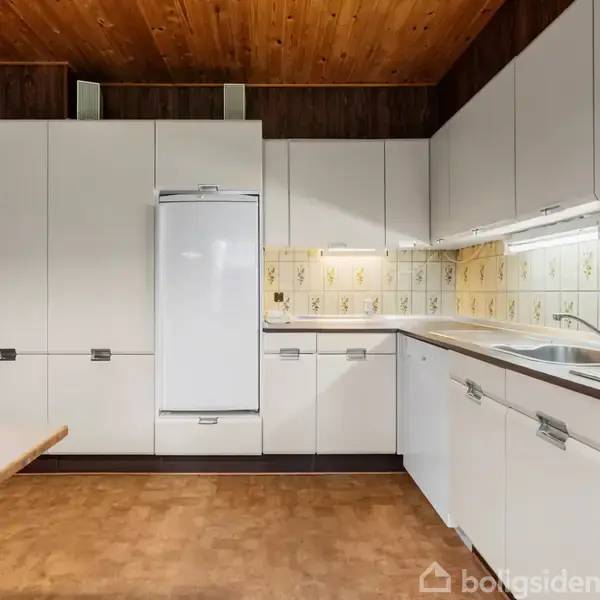 Køkken med hvide skabe i aktion, der står ordnet langs væggen; en opvaskemaskine og vask er indbygget. Fliser med mønstre dekorerer væggen under en træbeklædt loft. Ingen tekst bortset fra vandmærke.