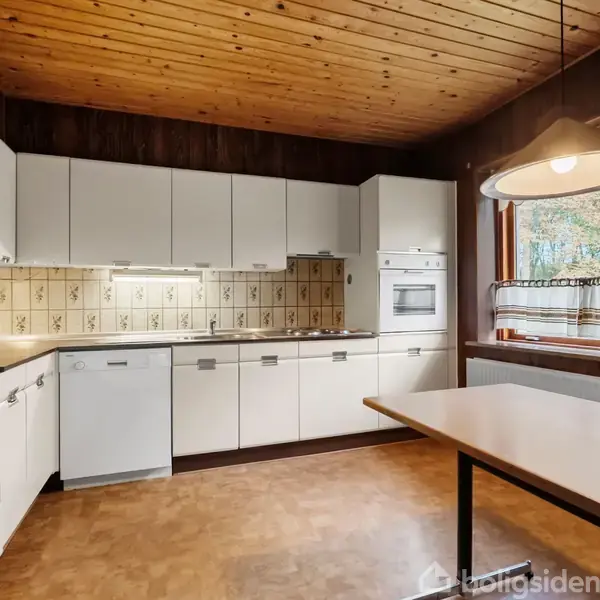 Køkken med hvide skabe arrangeret i en L-form. En indbygget ovn og opvaskemaskine er synlige. Træloft og parketgulv skaber en rustik atmosfære. Et stort vindue med udsigt udadtil.