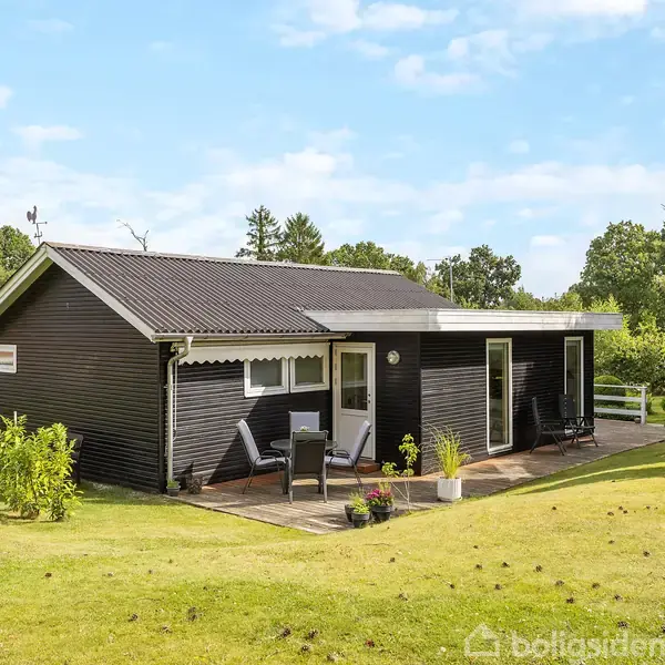 Et sortmalet sommerhus står stille på en grøn græsplæne omkranset af træer. Terrassen er dekoreret med planter i potter og havemøbler. Himmel er klar med få skyer.