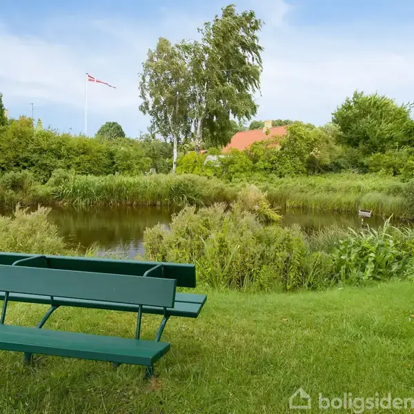 Grøn bænk står stille på en græsplæne. Foran den er en lille sø omgivet af buske og træer, med et hus og en flagstang i baggrunden.