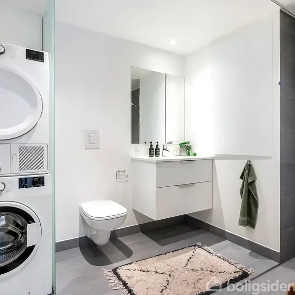 Vaskemaskine og tørretumbler står stablet. En hvid håndvask med spejl hænger over et skab. Ved siden af er et væghængt toilet. Håndklæde hænger til højre. Flisegulv og -vægge.
