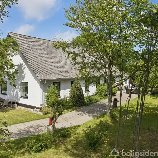 Hvidt hus med skråt tag står omgivet af træer og en velplejet have. Haven har en flisebelagt sti langs huset under en blå himmel med lette skyer.