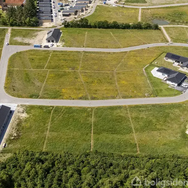 Grønne marker opdelt i byggegrunde omkranset af asfaltveje og nogle få huse. Skovområde i forgrunden og flere bygninger samt beplantning i baggrunden.