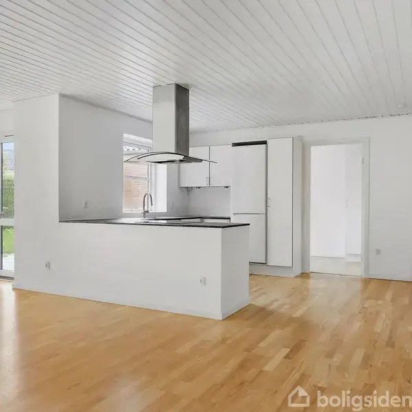 Køkken-alrum med hvidt loft og trægulv, centralt placeret kogeø med emhætte. Store vinduer på den ene væg giver udsigt til have og naturligt lys.