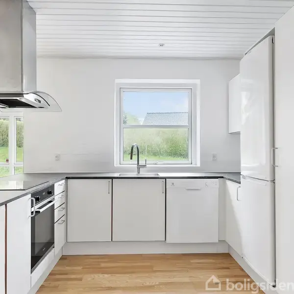 Et moderne køkken med hvide skabe og en sort bordplade, inkludert en ovn og komfur. Et stort vindue giver udsigt til en grøn have. Trægulv og hvidt loft.