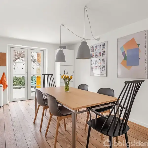 Bord med vase af gule blomster står i en spisestue med trægulv; omgivet af stole og lamper ovenover. Døren fører ud til en have. To vægplakater er synlige.