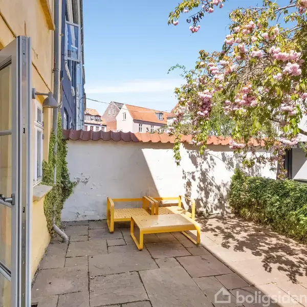 En gulmalet udendørs bænk står under et kirsebærtræ med lyserøde blomster i en flisebelagt gårdhave ved siden af en gul bygning med åbne glasdøre.