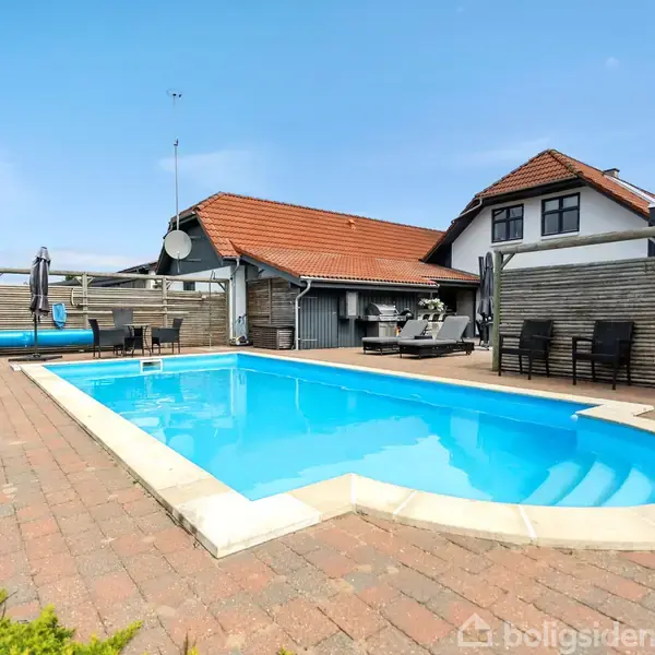 Swimmingpool med klart blåt vand omkranset af fliser. I baggrunden er et hus med rødt tegltag og havemøbler under en overdækket terrasse. Skyfri himmel i baggrunden.