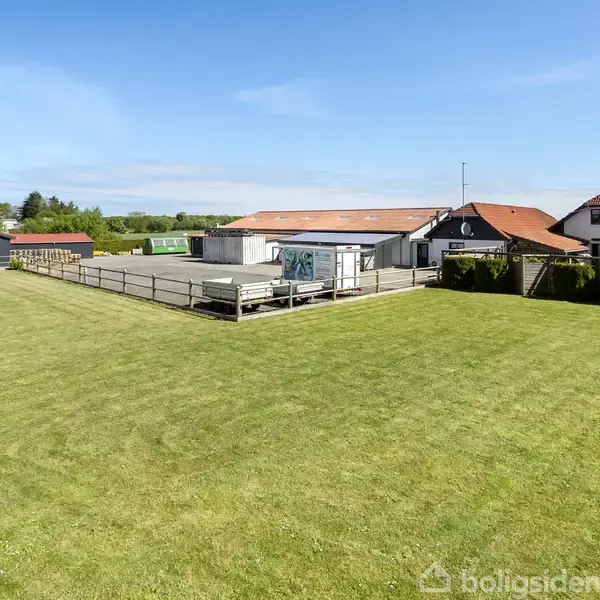 En grøn græsplæne strækker sig foran en gård med flere bygninger, herunder en med rødt tag. Himlen er blå, og landskabet er landligt.