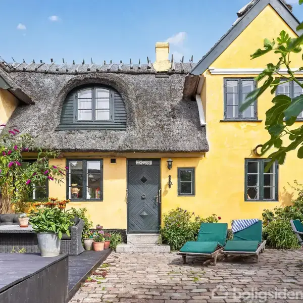 Rundt havebord omgivet af træstole med blå hynder står på en brostensbelagt gårdhave. Til højre en udendørs kamin med murstenstag, omkranset af grønne planter mod en gul mur.