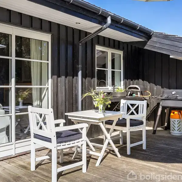 Terrasse med hvidt cafésæt, prydet af blomster i vase, foran sortmalet træbygning. Gasgrill står til højre. Sollys skaber en lys og indbydende atmosfære.