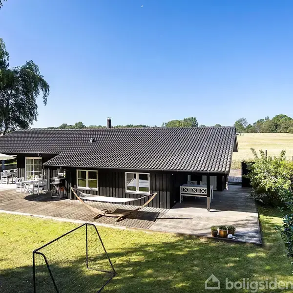 Sort træhus med terrasse og hængekøje, omgivet af grønt græs og træer. Huset ligger i et åbent, landligt område med en mark i baggrunden under en klar blå himmel.