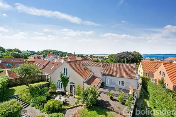 Hus med rødt tegltag står i en grøn have. Baggrunden viser flere huse og udsigt til vandet under en blå himmel med spredte skyer.