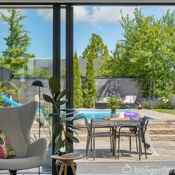 En grå lænestol med farverig pude står ved siden af et plantebord; udenfor ses terrasse med spisebord, stole, swimmingpool og grønne planter under blå himmel.
