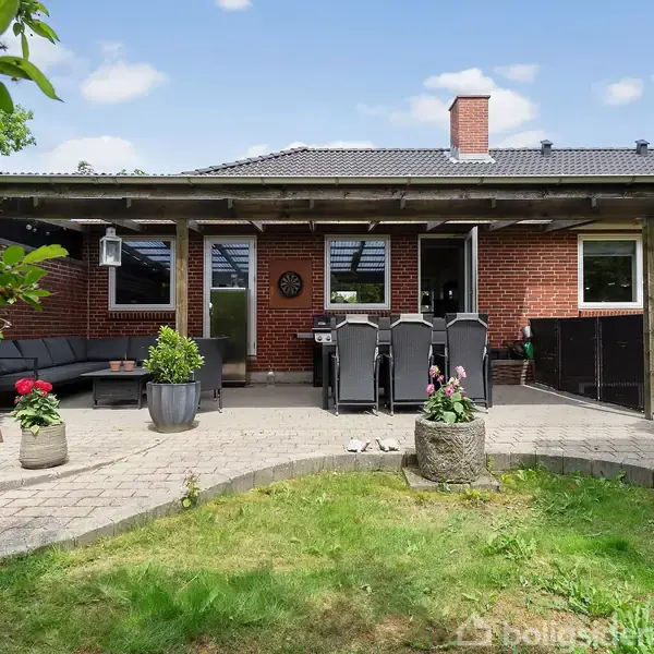 Terrassen har grå fliser og en pergola, der dækker siddeområdet med havemøbler. Omkring er der grønne græsplæner og træer. En rød murstensvæg udgør en del af huset.