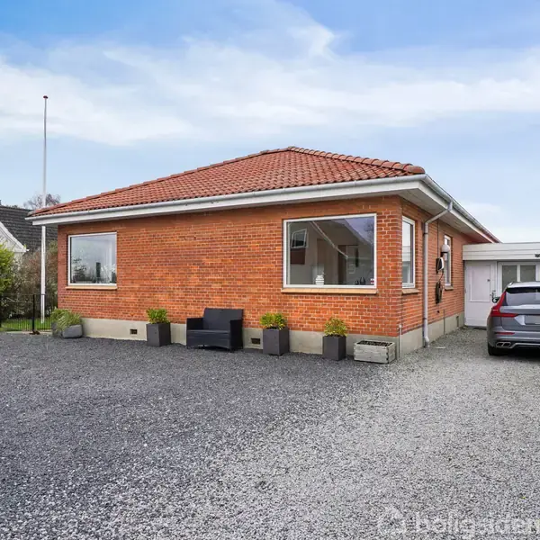 Rødt murstenshus med rødt tegltag, parkeret sort bil ved siden af. Sort bænk og blomsterkasser foran huset, omgivet af grusplads og hæk. Klar, blå himmel i baggrunden.