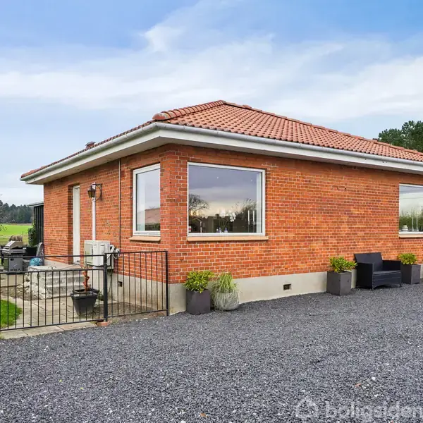 Rødt murstenshus med brunt tegltag står på en grusbelagt indkørsel. Store vinduer reflekterer den omkringliggende grønne græsplæne og træer, hvilket skaber en fredelig, landlig atmosfære.