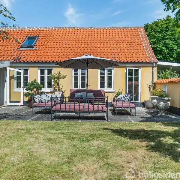 Gulmalet hus med rødt tegltag står i solskin. Terrasse foran huset med havemøbler og stor parasol. Græsplæne og træer omgiver området.