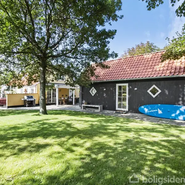 Et sort træhus står stille ved siden af en stor grøn græsplæne. Et surfbræt læner mod husets ydervæg. En terrasse med møbler befinder sig til venstre.