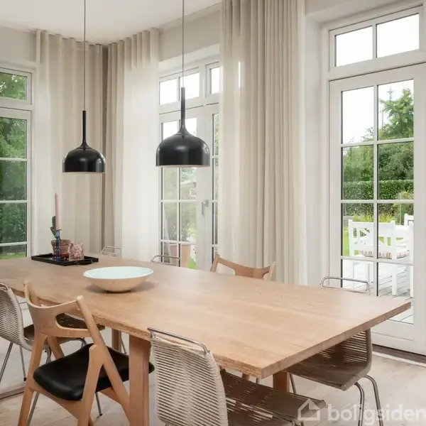 Et langt træbord står i en lys spisestue med moderne stole omkring. To sorte pendellamper hænger over bordet. Store vinduer og glasdøre åbner til en terrasse med grønne omgivelser.
