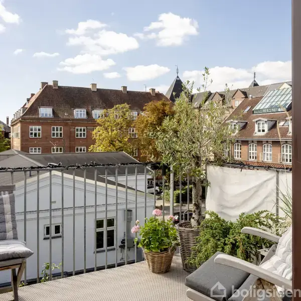 En altan med planter i krukker set over en gård. I baggrunden ses røde murstensbygninger med hvide vinduer og tage under en blå himmel med hvide skyer.