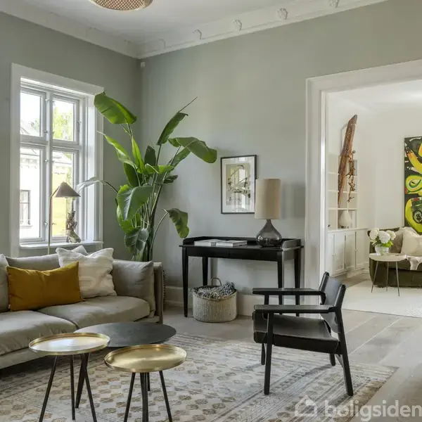 Stue indrettet med grå sofa og gul pude ved siden af grøn plante. To små runde borde foran sofaen. Lys farve på væggene, store vinduer, kunstværk hænger i naborummet.