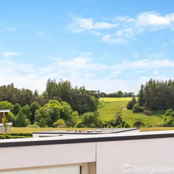 En grøn mark med bakker og træer ses i baggrunden, omgivet af skov, under en klar blå himmel. En murstensbygning er delvist synlig til venstre.