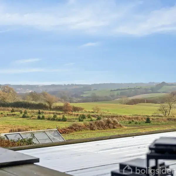 Landskabsudsigt viser et grønt, bakket terræn med marker og spredte træer under en klar blå himmel. Et glasværn og terrassekant ses i forgrunden. Ingen tekst til stede.