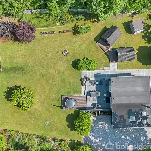 En velholdt have med grøn græsplæne omgiver et moderne hus. Der er tre små udhuse og et cirkulært bålsted i græsset. Omkranset af træer og buske.