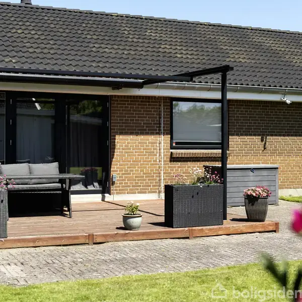 En enkelthus af mursten med fladt tag står på en stor, velplejet græsplæne. Huset har en terrasse med havemøbler, og træer omkranser ejendommen i baggrunden.