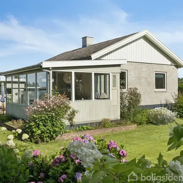 Et hus med glasveranda står i en frodig have med farverige blomster. Huset har gråt murværk og hvidt træværk. Himlen er klar og blå.