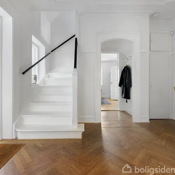 En trappe fører opad langs en hvidmalet væg med håndliste. Indgangen til huset har trægulv, åbne døre viser flere rum. En jakke hænger ved døren, taske på panel.