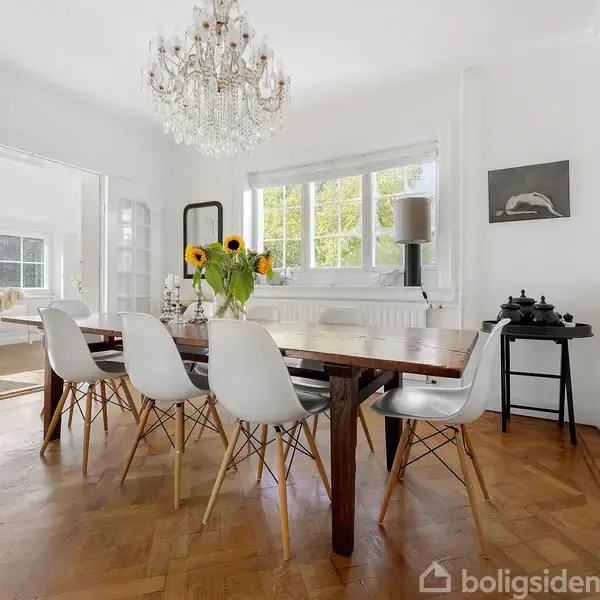 Spisebord med hvide stole omringet af lyse vægge og gulv i parket. En lysekrone hænger over bordet, hvor en vase med solsikker står. Rummet er lyst med store vinduer.