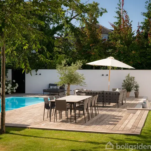 En træterrasse med spisebord og stole under en åben parasol, der står ved siden af en swimmingpool. Omkring er der grønne træer og en græsplæne i en privat baghave.