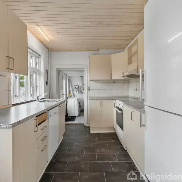 Et hvidt køkken med klinkegulv har moderne apparater såsom opvaskemaskine, komfur og stort køleskab. Lang bordplade strækker sig langs vinduerne, der giver naturligt lys.