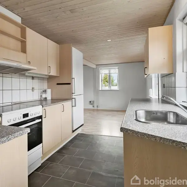 Køkken med grå flisegulv og lyse trækøkkenskabe, hvid ovn og emhætte. Køkkenet har en stor vask, gråt bordplade, og åbner mod en stue med vindue og lyst trægulv.