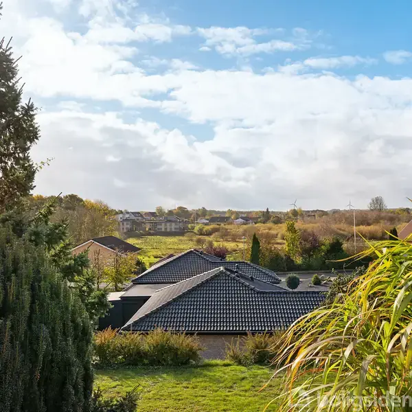Et hus med sort tag ligger stille i en grøn have med træer og buske. I baggrunden ses et åbent landskab med skyer på en blå himmel.