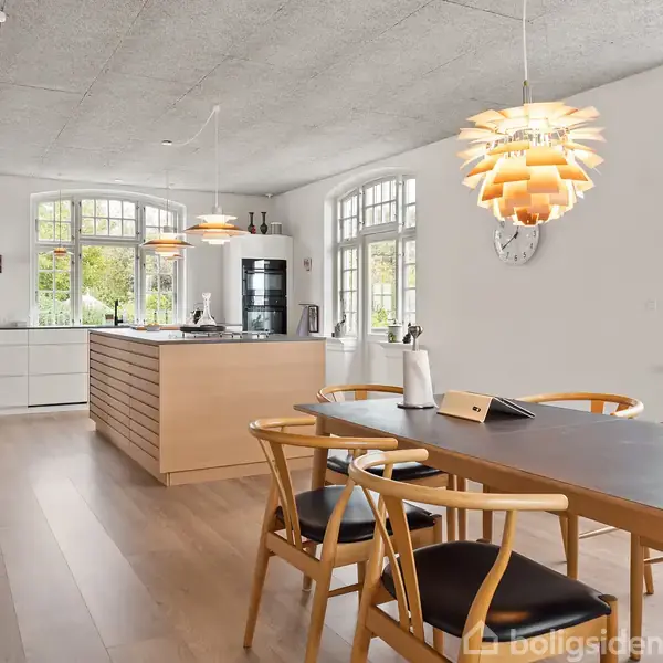 Køkken-alrum med træspisebord og stole, køkkenø i træ, hvide skabe. Hængelamper over bordet. Store vinduer slipper naturligt lys ind og planter ses udenfor.