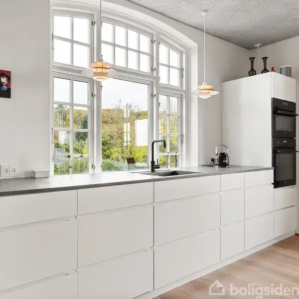 Hvidt køkken med håndvask og ovne i skabsvæg, set i dagslys fra store buede vinduer. Trænger til vedligeholdelse. To lamper hænger over bordpladen. Ingen tekst synlig.