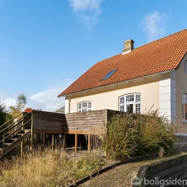 Hus med rødt tegltag og beige facade, omgivet af terrasse med trappe i en have. Solskin og blå himmel i baggrunden.