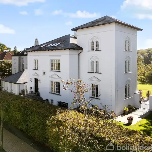 Hvid villa med tårnstruktur, står stille omgivet af frodig have og grønne hække i et forstadsområde med andre huse.