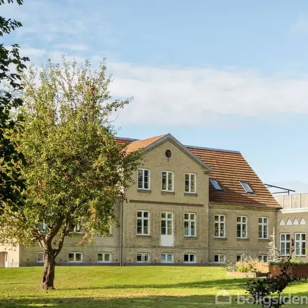 En stor, lys murstensbygning med rødt tegltag står på en solrig græsplæne. Omkranset af træer og en blå himmel, ser den ud som en del af et landligt landskab.