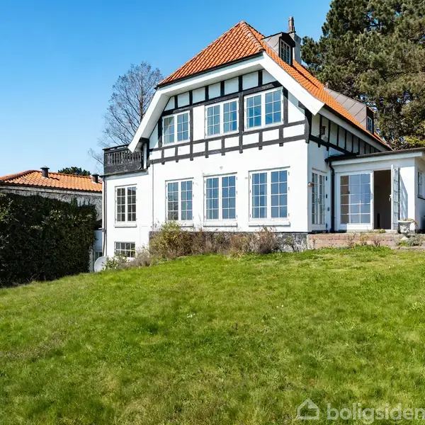 Hus med bindingsværk står på en grøn græsplæne i en have. Der er et orange tegltag og flere store vinduer. Baggrunden viser træer og en klar blå himmel.