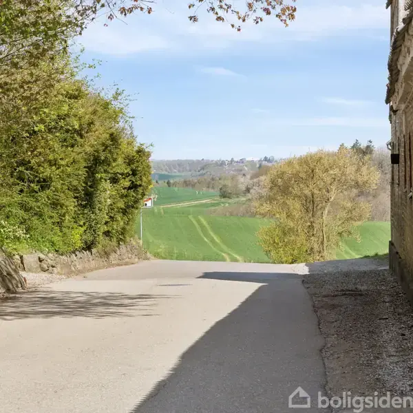 Vej fører nedad, flankeret af murstensbygning til højre og frodig vegetation til venstre. I baggrunden ses grønne marker under en klar, blå himmel.