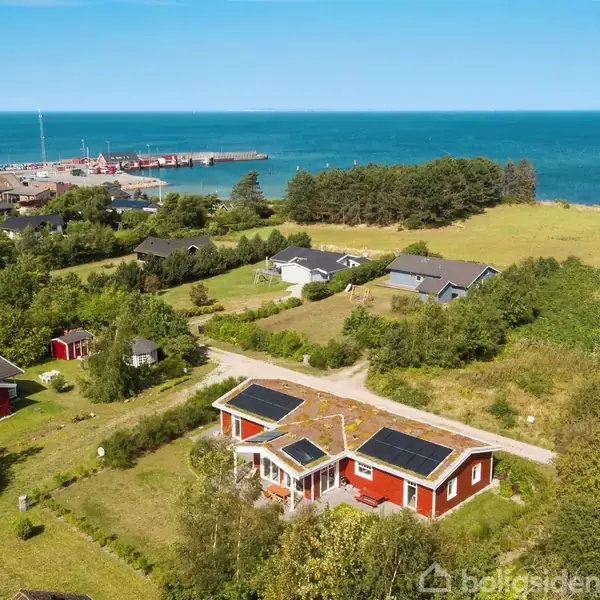 Rødt hus med solpaneler står stille i grønt landskab nær kysten. Omringet af andre huse, skov og strand, med udsigt til havn og hav.