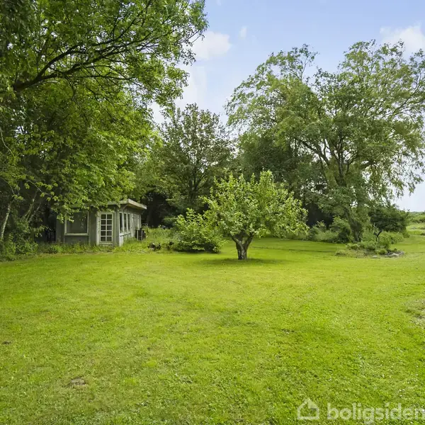 Et træhus står omgivet af en frodig have med en græsplæne og store træer. I baggrunden ses en åben mark under en klar blå himmel.